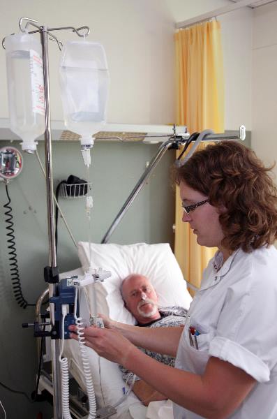 \"verpleegkundige Afbeelding van een verpleegkundige die het infuus controleert van iemand die in een ziekenhuisbed ligt. Hij is ondertussen in gesprek met de patië®´.\"